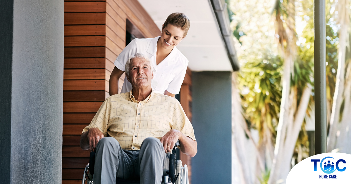 A caregiver brings an elderly man in a wheelchair out of a building, demonstrating a hospital discharge.