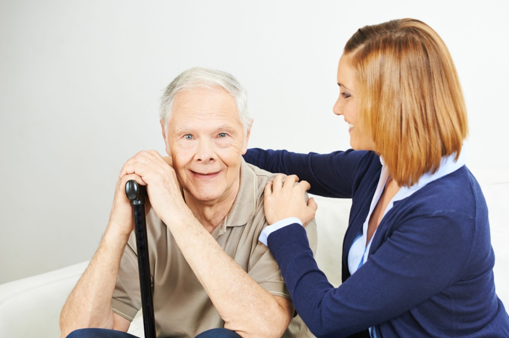A cheerfull care giver attends to and elderly man sitting on a couch