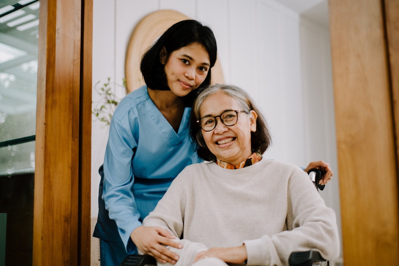 A smiling elderly woman in a wheelchair is being gently assisted by a young female caregiver in blue scrubs. They are standing at the entrance of a cozy, well-lit home. This image represents compassionate senior care in San Gabriel, showcasing warmth, support, and a trusting caregiver-patient relationship.
